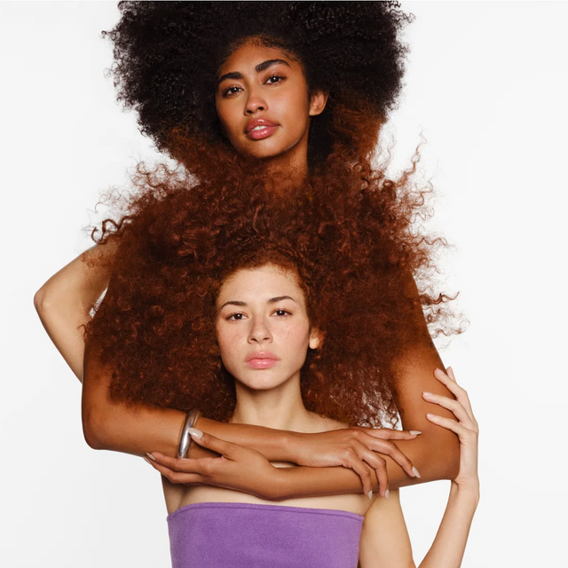 Two women with voluminous curly hair pose together against a white background. One stands behind, resting her arms gently on the shoulders of the other, who wears a purple towel and gazes directly at the camera. Their healthy curls showcase the results of dedicated scalp care with Isima’s Iconic Scalp System, revealing noticeable hair growth that can be achieved in as little as 90 days.