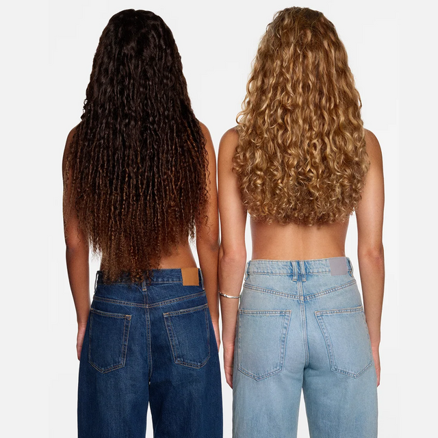 Two women stand side by side, backs to the camera, wearing blue jeans and no shirts. One shows off long dark curls while the other’s slightly shorter ringlets are a lighter shade—both revealing hair that looks vibrant and utterly free from damage. Against a simple white background, their striking silhouettes radiate confidence as they model beautiful, well-cared-for curls—each enhanced by isima’s Shakira’s Súperbomba Set.