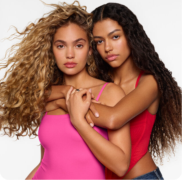 Two women with long, curly hair pose closely together against a white background. One wears a bright pink top, and the other a red top, with their arms gently wrapped around each other.
